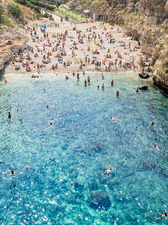 A vertical image of the italian coast at Polignano al Mare featuring beach goers in the ocean and on the sand