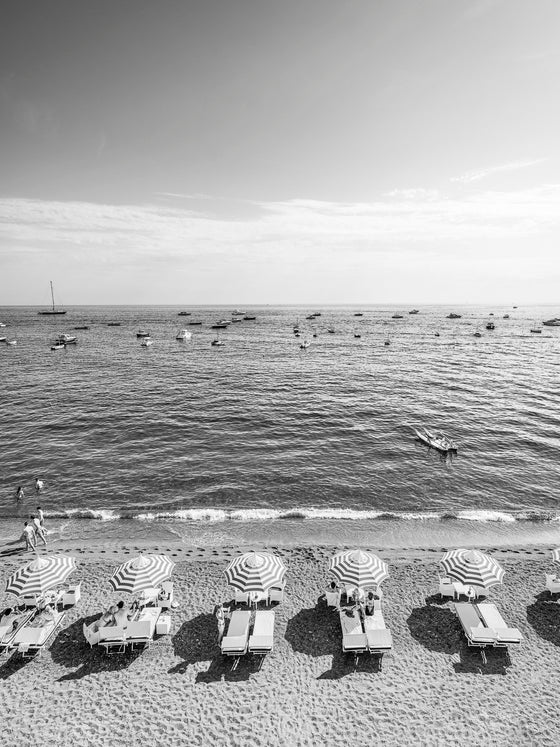 Black and white photo of five striped beach umbrellas with sun beds and the mediterranean ocean with boats and the horizon and sky in the background.