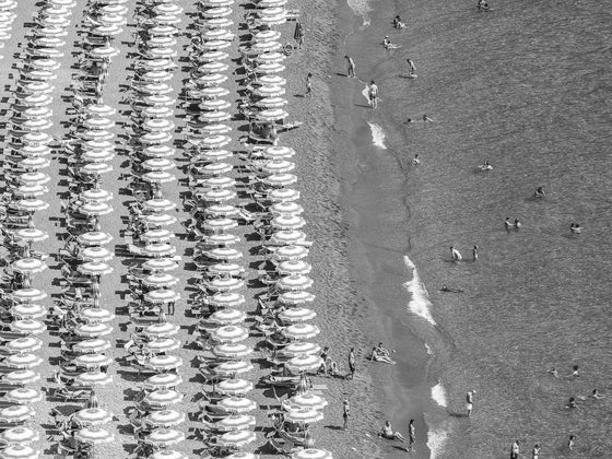 Black and white beach umbrellas on the Positano Beach on the Amalfi Coast in Italy on the left half and water with a few people to the right half in the summertime.