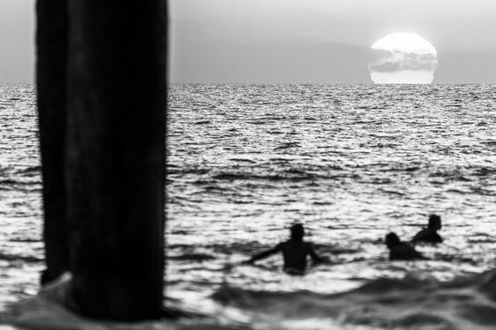 Three surfers in Manhattan Beach, CA enjoying the sunset from the water. The sky is orange and the large yellow sun can be seen starting its descent below the horizon. This version of the photo is in black and white.