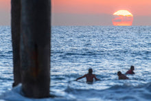  Three surfers in Manhattan Beach, CA enjoying the sunset from the water. The sky is orange and the large yellow sun can be seen starting its descent below the horizon.