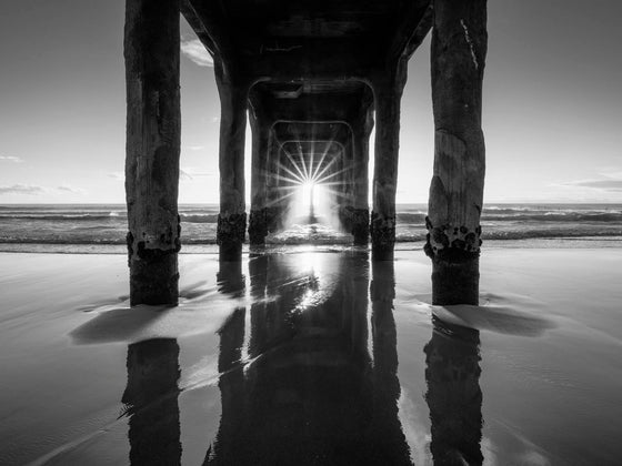 Under the Manhattan Beach Pier at low tide with the sun streaming in and mist and a starburst, during the annual pierhenge day when the setting sun lines up directly down the middle of the Manhattan Beach Pier, in black and white