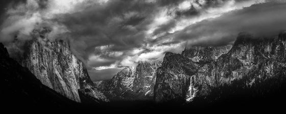 A panoramic image of the Yosemite clearing during sunset, featuring a waterfall and clouds, in black and white